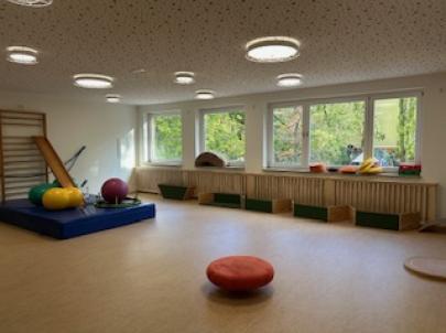 Ein heller Raum mit großen Fenstern, der mit verschiedenen Sportgeräten ausgestattet ist. Auf dem Boden liegen ein runder Sitzpuff und mehrere Bälle.