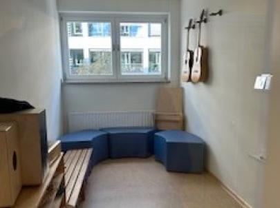  Ein Raum mit einer Holzbank und zwei Gitarren an der Wand. Der Raum ist hell und freundlich gestaltet, mit einem Fenster, das Tageslicht hereinlässt.
