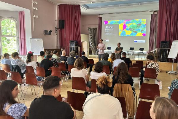 Zwei erwachsene Personen stehen in einer Aula. Im Hintergund ist eine bunte Präsentation zu sehen. Im Vordergrund sieht man das Publikum, ca. 30 Personen, die auf Stühlen sitzen.