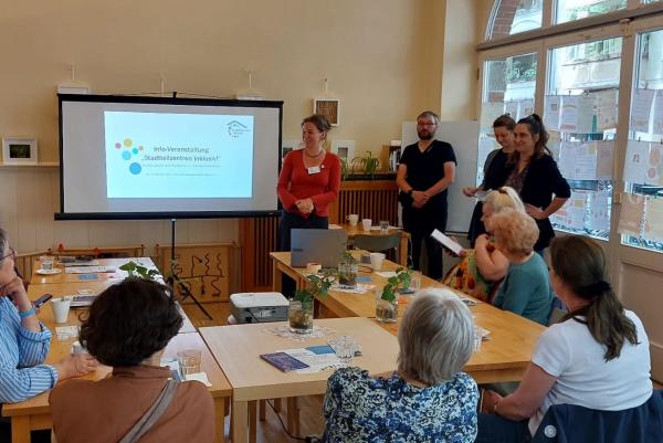 Vier erwachsene Menschen, stehen neben einer Leinwand, auf der eine Präsentation zu sehen ist. Dort steht „Info-Veranstaltung, Stadteilzentren inklusiv!“. Vor der Leinwand steht eine U-förmige Tischgruppe, an der sechs weitere erwachsene Menschen sitzen und aufmerksam die Vortragenden und die Präsentation betrachten.