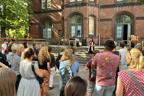 Direktorin Henriette Harms hält eine Rede. Sie steht auf einer Treppe und spricht zum Publikum. Im Hintergrund sieht man ein Backsteingebäude. Im Vordergrund stehen Menschen und der Rede hören zu. 
