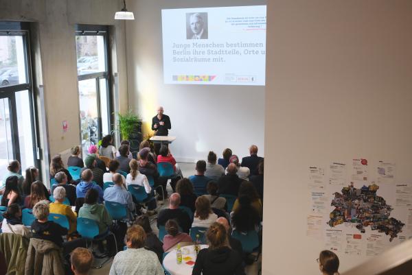 Eine Gruppe von Menschen sitzt in einem Raum und hört einem Vortrag zu. Ein Mann steht am Rednerpult und spricht, während im Hintergrund eine Präsentation an einer Wand gezeigt wird.