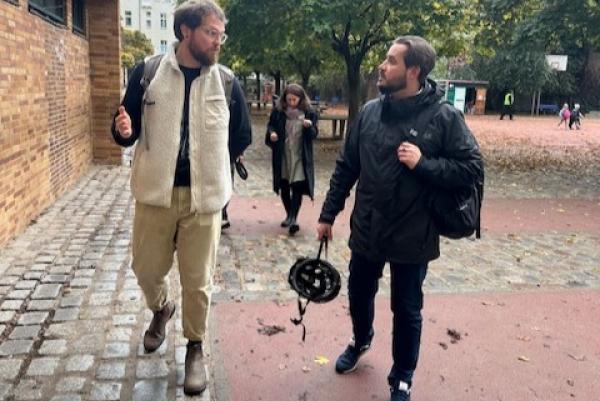 Eine Aufnahme von zwei Männern, die auf einem gepflasterten Weg in einem Hof gehen. Der Mann links trägt eine helle, gefütterte Weste, eine schwarze Hose und hat eine Brille auf. Er gestikuliert mit der linken Hand, während er spricht. Der Mann rechts trägt eine schwarze Jacke und hält einen Fahrradhelm in der rechten Hand. Im Hintergrund ist eine Frau zu sehen, die in einer dunklen Jacke gekleidet ist und ebenfalls in Richtung der beiden Männer geht. Der Hof hat Bäume und einen Spielplatz im Hintergrund.