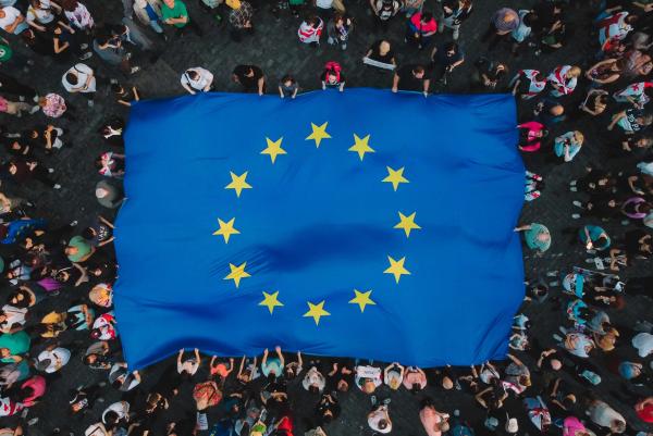 Vogelperspektive auf eine Menschenmenge, die eine große europäische Flagge hält. Die Flagge ist blau mit einem gelben Kreis aus zwölf Sternen in der Mitte. Um die Flagge herum sind viele Menschen zu sehen, die unterschiedliche Kleidung tragen Der Boden ist mit dunklen Steinen gepflastert. Die Szene wirkt lebhaft.