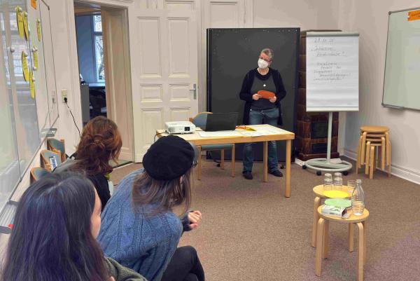 Eine Person steht an einem Tisch und hält einen orangefarbenen Zettel. Im Hintergrund sitzen drei Personen, die aufmerksam zuhören. An einer Wand steht ein Flipchart mit Notizen.