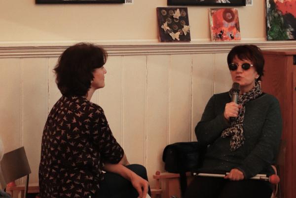 Zwei Frauen sitzen sich gegenüber. Die eine Frau hält einen Zettel in der Hand, die andere Frau hält ein Mikrophon und spricht. Sie trägt eine Sonnenbrille.