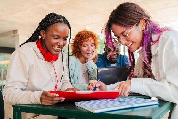 4 junge Personen sitzen an einem Tisch, lächeln und Arbeiten gemeinsam mit Laptop und Arbeitsunterlagen.