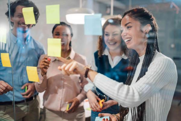 Eine Gruppe von vier Personen steht an einer Glaswand und organisiert bunte Haftnotizen. Im Vordergrund zeigt eine Frau mit geflochtenen Haaren und weißem Pullover auf eine Notiz; weitere Personen (zwei Frauen, ein Mann) beobachten und halten Stifte oder weitere Notizzettel. Die Szene wirkt wie ein Team-Workshop oder Brainstorming in einem modernen Büro.