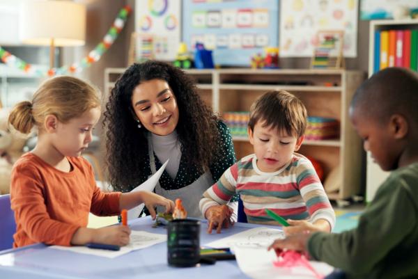 Eine Erzieherin sitzt mit vier Kindern an einem Tisch in einem bunten Kindergartenraum; sie malen und basteln gemeinsam.