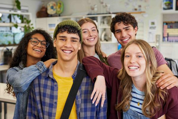 Fünf lächelnde Jugendliche stehen eng beieinander in einem Klassenraum, Arme umeinander gelegt, und blicken fröhlich in die Kamera.