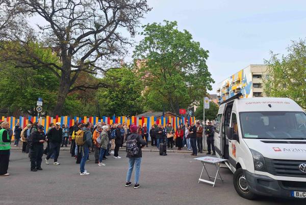 Straßenszene mit einer Menschenmenge vor bunt gestreiftem Zaun; rechts steht ein weißer Caritas‑Arztmobil‑Van mit offenem Schiebetür und Klapptisch. Bäume und Wohnhäuser im Hintergrund, Versammlung im Freien.