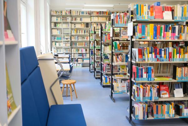 Helle Bibliothek mit Reihen voll bestückter Regale rechts, Arbeitsplätzen und blauen Sitzbänken links; ruhiger, aufgeräumter Lesebereich.