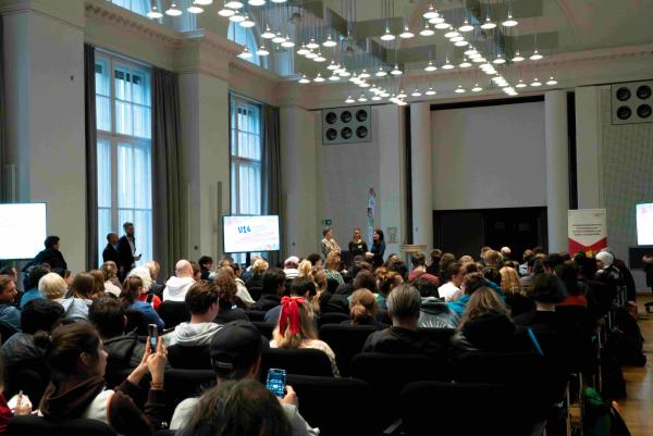 Großer Saal mit vielen sitzenden Teilnehmenden, Blick nach vorn zur Bühne. Drei Personen stehen vorne und sprechen; seitlich stehen Bildschirme mit der Aufschrift „U16“ und Veranstaltungsinfos. Hohes Deckenlicht, große Fenster, Konferenzatmosphäre.