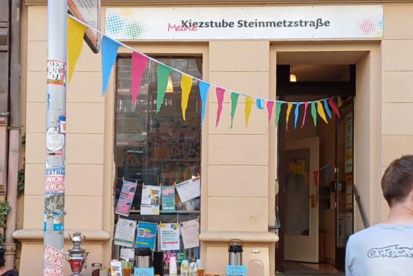 Eingang der „Kiezstube Steinmetzstraße“ mit bunter Wimpelkette; vor dem Schaufenster stehen Thermoskannen und Infomaterial, im Fenster hängen Flyer.