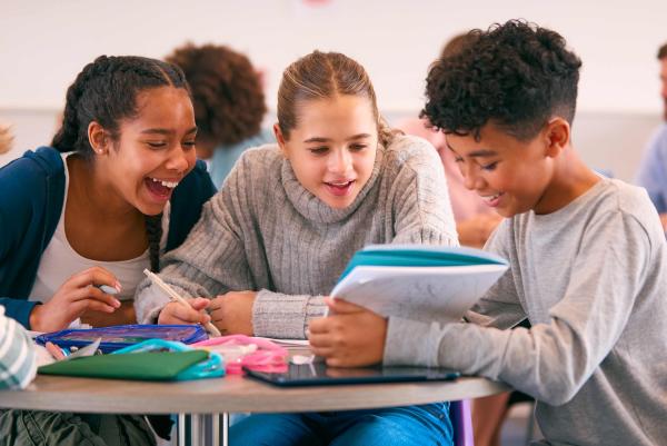 Vier Schulkinder sitzen lächelnd an einem Tisch und schauen gemeinsam in ein Heft; sie arbeiten mit Stiften und bunten Mappen; im Hintergrund unscharf weitere Personen im Klassenraum.