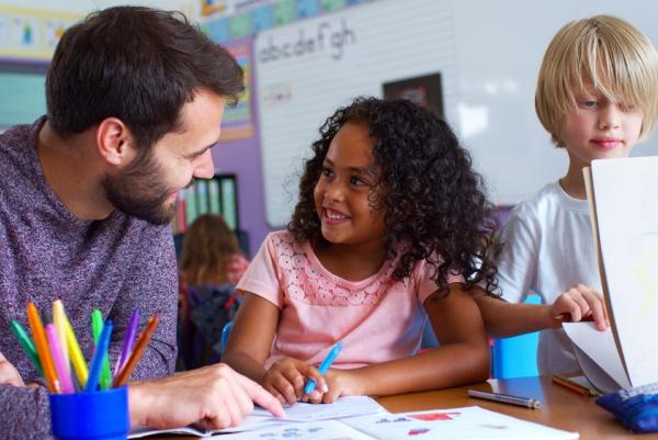Lehrer und zwei Kinder (Mädchen und Junge) arbeiten gemeinsam an einem Tisch; das Mädchen schreibt, der Junge hält einen Schreibblock; im Hintergrund ein Klassenzimmer mit weiteren Kindern.