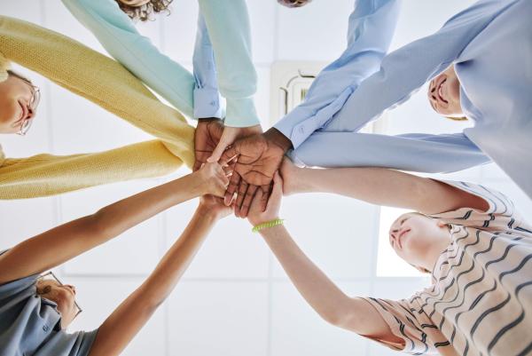 Fünf Personen stehen im Kreis und legen ihre Hände übereinander in die Mitte. Die Hände sind unterschiedlich und symbolisieren Gemeinschaft und Zusammenhalt. Im Hintergrund sind lächelnde Gesichter zu erkennen.