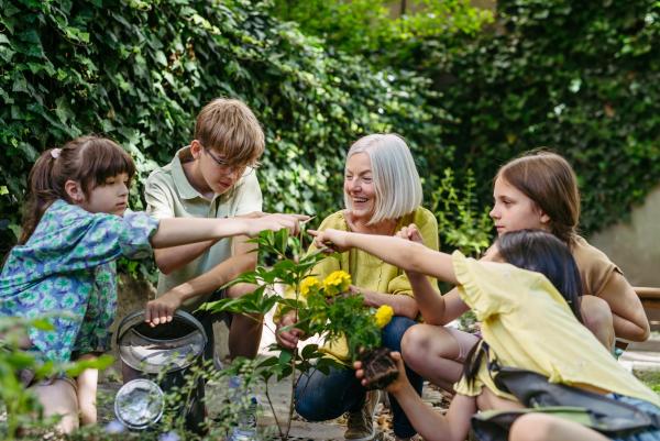 Eine Frau mit weißem Haar und gelbem Oberteil sitzt lachend im Freien und zeigt gemeinsam mit vier Kindern auf eine blühende Pflanze mit gelben Blüten. Im Hintergrund eine dichte grüne Hecke; eines der Kinder hält eine Gießkanne.