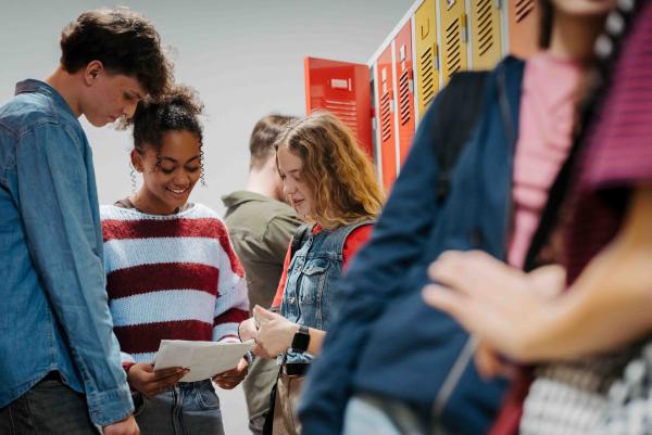 Gruppe von Jugendlichen steht im Schulflur vor bunten Schließfächern; zwei Mädchen lesen gemeinsam ein Blatt Papier und lächeln, ein Junge und weitere Schüler*innen stehen in der Nähe.