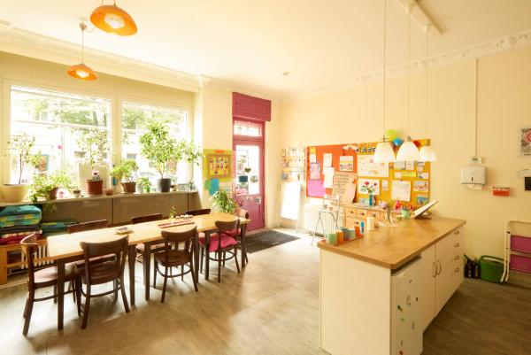 Freundlicher Caféraum mit großen Fensterfronten, vielen Pflanzen, langem Holztisch mit Stühlen und Infowand; daneben die Theke mit bunten Bechern.