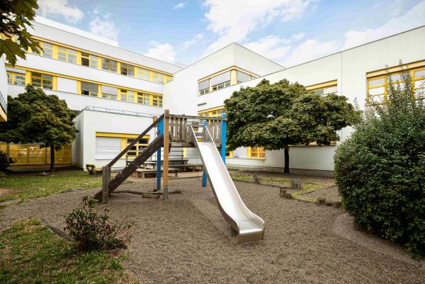 Innenhof‑Spielplatz vor weißem Gebäude mit gelben Fenstern: Kletterturm mit Rutsche, Sitzbänke und Bäume rundherum.