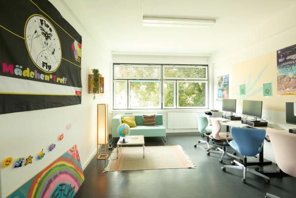 Freundlicher Computer- und Treffraum mit pastellfarbener Couch und Teppich, mehreren PC-Arbeitsplätzen an der rechten Wand und großen Fenstern mit Blick ins Grüne.