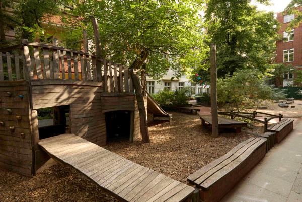 Schattiger Kita‑Hof mit großen Spielplatz aus Holz, Sitzbänken, Bäumen und Sträuchern.