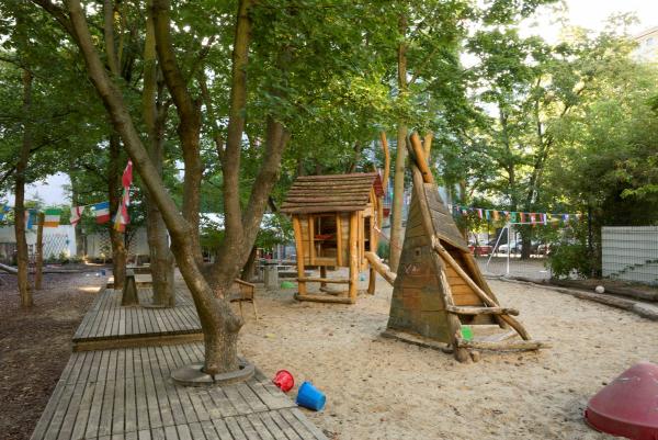 Schattiger Kita‑Spielplatz mit vielen Bäumen: Sandfläche mit Holzhäuschen und Spielzeugen; im Hintergrund bunte Wimpelkette und ein Fußballtor.