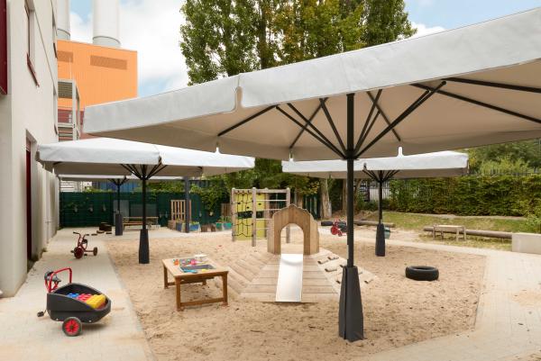 Heller Sandspielbereich mit großen Sonnenschirmen, kleinem Kletter‑ und Rutschhügel, Werkbanktisch und Dreirädern; Hochbeete und Sitzbänke am Rand.