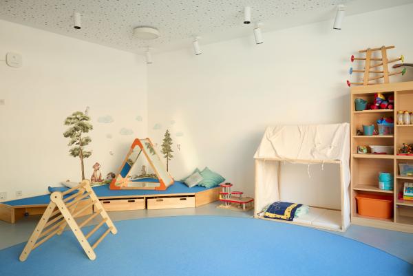 Heller Spielraum im Nestbereich mit großem blauem Teppichkreis, hinten eine niedrige gepolsterte Ecke mit Wandmalerei und Spielzeug; rechts ein kleines Stoffzelt mit Matten und Decken sowie ein offenes Regal mit sortierten Spielmaterialien.
