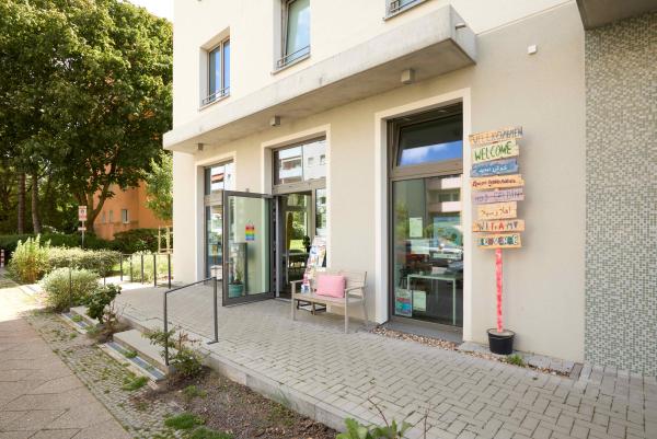 Eingang eines modernen Nachbarschaftszentrums mit beiger Fassade, Glastür, Holzbank mit rosa Kissen und mehrsprachigem Willkommensschild an der Wand. Gepflasterte Terrasse mit Grünflächen und Bäumen im Hintergrund.