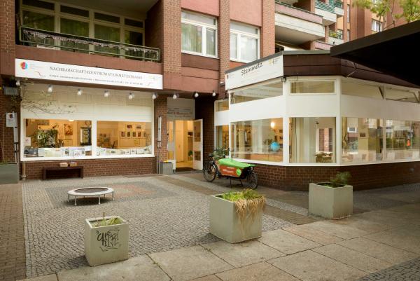  Außenansicht des Nachbarschaftszentrums in einem roten Klinkerbau. Große Schaufenster geben den Blick in helle Räume frei; vor dem Eckfenster steht ein grünes Lastenrad. Davor ein kleiner gepflasterter Platz mit Pflanzkübeln.