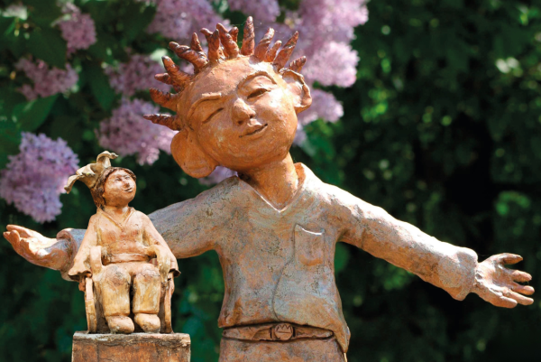  Kinderkönig*innen: Eine Tonfigur eines Jungen mit ausgebreiteten Armen vor blühenden Sträuchern; links neben ihm sitzt eine zweite Figur im Rollstuhl.