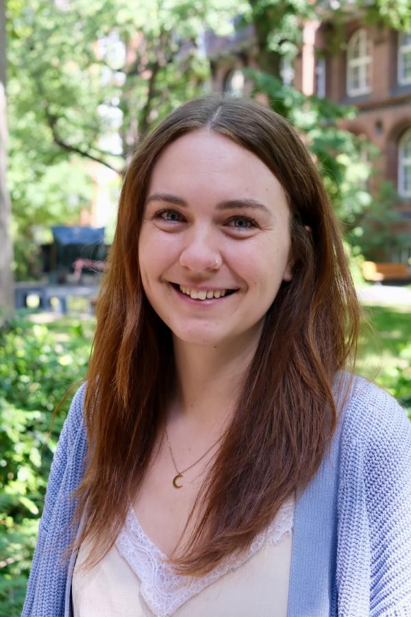 Eine Frau mit langen braunen Haaren lächelt in die Kamera. Sie trägt eine hellblaue Strickjacke und ein beiges Top. Im Hintergrund sieht man grüne Bäume und ein Backsteingebäude. 