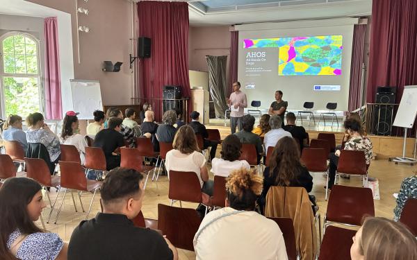 Zwei erwachsene Personen stehen in einer Aula. Im Hintergund ist eine bunte Präsentation zu sehen. Im Vordergrund sieht man das Publikum, ca. 30 Personen, die auf Stühlen sitzen.