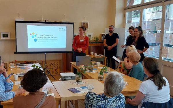 Vier erwachsene Menschen, stehen neben einer Leinwand, auf der eine Präsentation zu sehen ist. Dort steht „Info-Veranstaltung, Stadteilzentren inklusiv!“. Vor der Leinwand steht eine U-förmige Tischgruppe, an der sechs weitere erwachsene Menschen sitzen und aufmerksam die Vortragenden und die Präsentation betrachten.