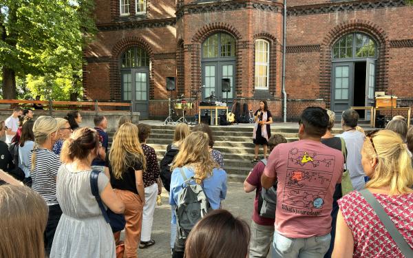 Direktorin Henriette Harms hält eine Rede. Sie steht auf einer Treppe und spricht zum Publikum. Im Hintergrund sieht man ein Backsteingebäude. Im Vordergrund stehen Menschen und der Rede hören zu. 