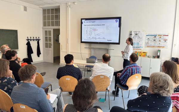 Dr. Marlies Kroetsch hält einen Vortrag. Im Hintergrund sieht man eine Präsentation mit einer Grafik. Im Vordergrund sitzen ca. 15 Personen und hören aufmerksam zu. 