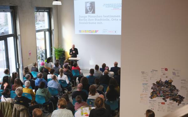 Eine Gruppe von Menschen sitzt in einem Raum und hört einem Vortrag zu. Ein Mann steht am Rednerpult und spricht, während im Hintergrund eine Präsentation an einer Wand gezeigt wird.