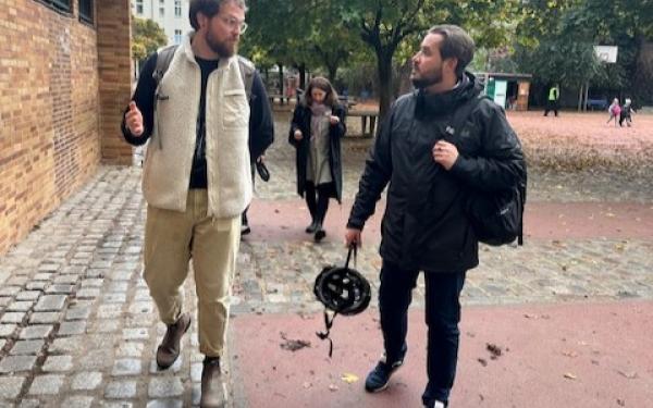 Eine Aufnahme von zwei Männern, die auf einem gepflasterten Weg in einem Hof gehen. Der Mann links trägt eine helle, gefütterte Weste, eine schwarze Hose und hat eine Brille auf. Er gestikuliert mit der linken Hand, während er spricht. Der Mann rechts trägt eine schwarze Jacke und hält einen Fahrradhelm in der rechten Hand. Im Hintergrund ist eine Frau zu sehen, die in einer dunklen Jacke gekleidet ist und ebenfalls in Richtung der beiden Männer geht. Der Hof hat Bäume und einen Spielplatz im Hintergrund.