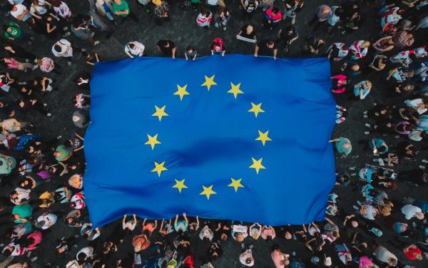 Vogelperspektive auf eine Menschenmenge, die eine große europäische Flagge hält. Die Flagge ist blau mit einem gelben Kreis aus zwölf Sternen in der Mitte. Um die Flagge herum sind viele Menschen zu sehen, die unterschiedliche Kleidung tragen Der Boden ist mit dunklen Steinen gepflastert. Die Szene wirkt lebhaft.