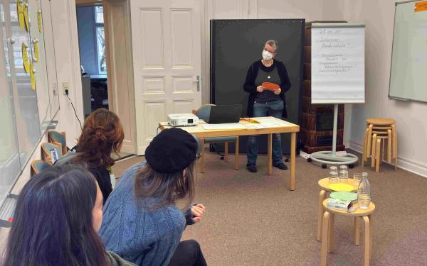 Eine Person steht an einem Tisch und hält einen orangefarbenen Zettel. Im Hintergrund sitzen drei Personen, die aufmerksam zuhören. An einer Wand steht ein Flipchart mit Notizen.