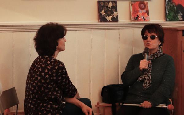 Zwei Frauen sitzen sich gegenüber. Die eine Frau hält einen Zettel in der Hand, die andere Frau hält ein Mikrophon und spricht. Sie trägt eine Sonnenbrille.