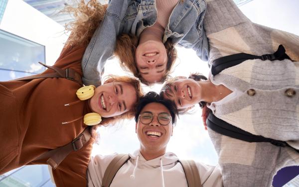 Vier junge Menschen bilden einen Kreis, schauen lachend nach unten in die Kamera und legen die Arme umeinander, mit Gebäuden und Himmel im Hintergrund.