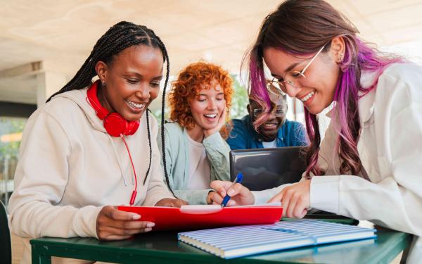 4 junge Personen sitzen an einem Tisch, lächeln und Arbeiten gemeinsam mit Laptop und Arbeitsunterlagen.