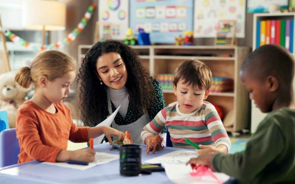 Eine Erzieherin sitzt mit vier Kindern an einem Tisch in einem bunten Kindergartenraum; sie malen und basteln gemeinsam.