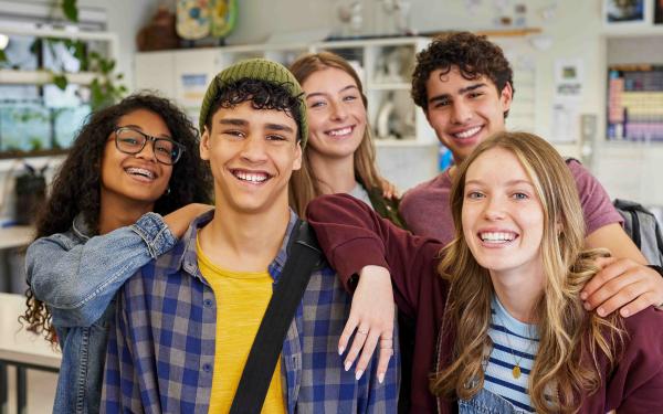 Fünf lächelnde Jugendliche stehen eng beieinander in einem Klassenraum, Arme umeinander gelegt, und blicken fröhlich in die Kamera.