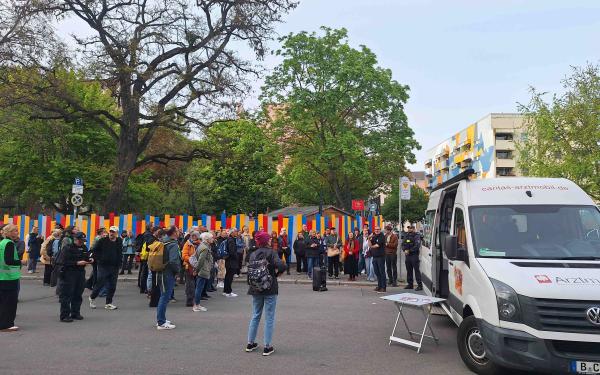 Straßenszene mit einer Menschenmenge vor bunt gestreiftem Zaun; rechts steht ein weißer Caritas‑Arztmobil‑Van mit offenem Schiebetür und Klapptisch. Bäume und Wohnhäuser im Hintergrund, Versammlung im Freien.