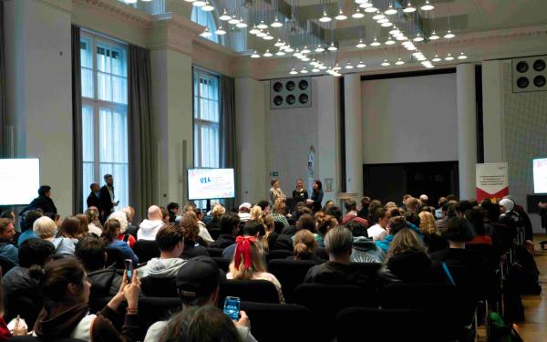 Großer Saal mit vielen sitzenden Teilnehmenden, Blick nach vorn zur Bühne. Drei Personen stehen vorne und sprechen; seitlich stehen Bildschirme mit der Aufschrift „U16“ und Veranstaltungsinfos. Hohes Deckenlicht, große Fenster, Konferenzatmosphäre.