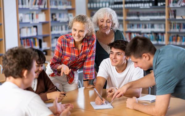 Gruppe Jugendlicher und eine ältere Lehrperson arbeiten lachend an einem Tisch in einer Bibliothek; sie diskutieren, schreiben Notizen und zeigen auf ein Blatt Papier.